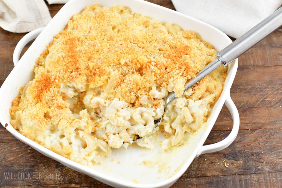 Crab mac and cheese in a baking dish with a spoon scooping some out on a wood table.