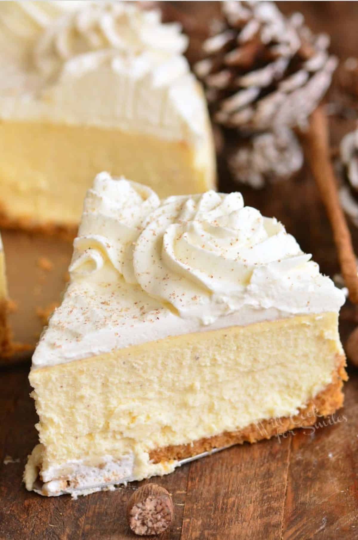 slice of eggnog cheesecake on a wood surface with the rest of the cheesecake and pinecones in the background.