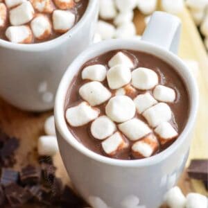 two mugs of hot chocolate with marshmallows in on top and sliced chocolate and marshmallows aground the bottom.