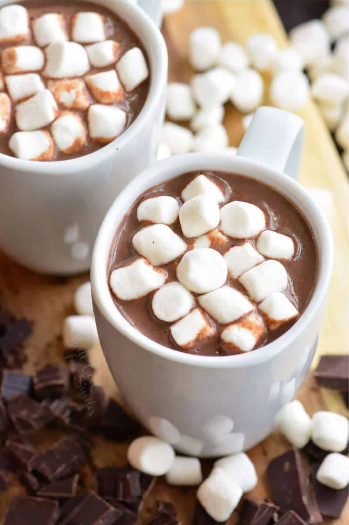 two mugs of hot chocolate with marshmallows in on top and sliced chocolate and marshmallows aground the bottom.