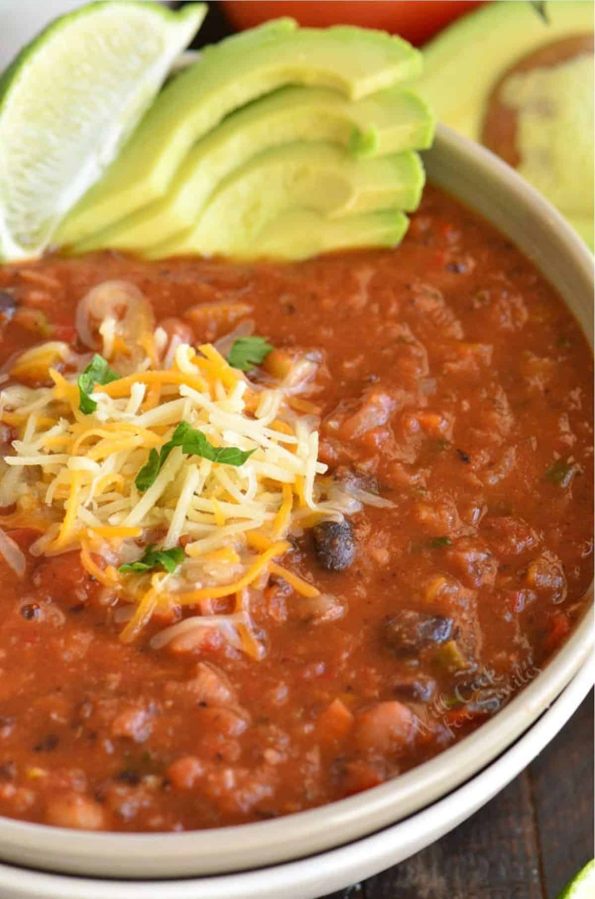 vegetarian chili in a bowl with a shredded cheese, a lime wedge, and sliced avocado for garnish.