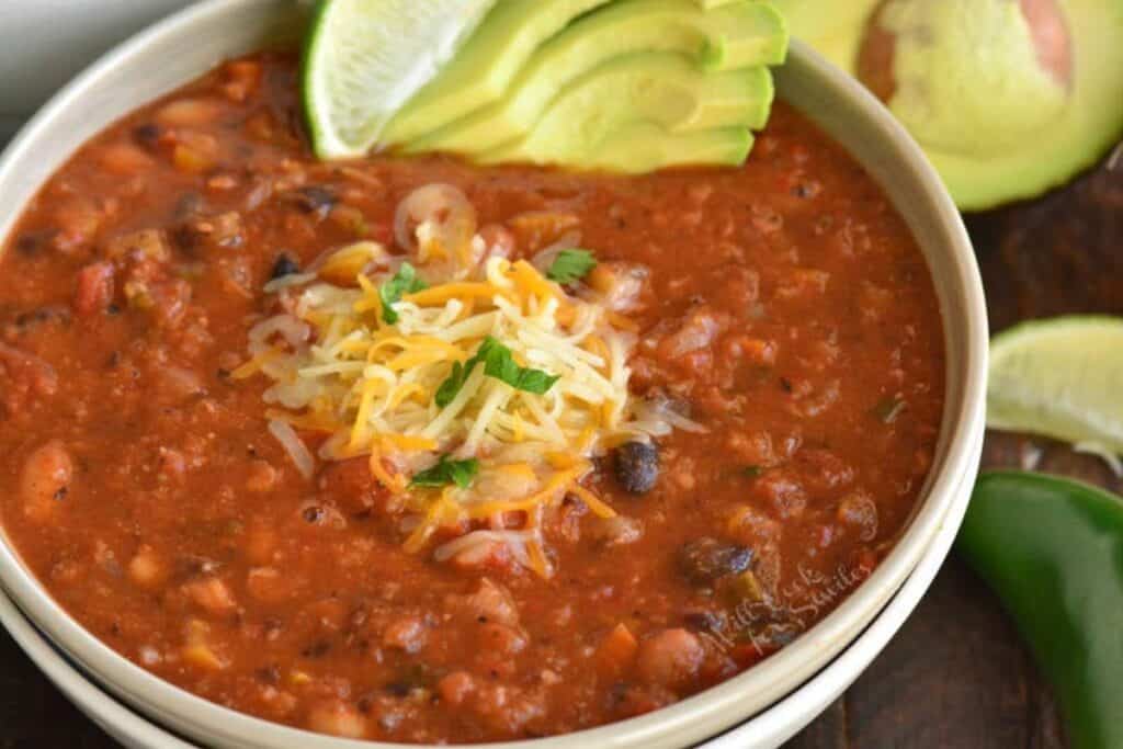 A bowl of hearty chili topped with shredded cheese and herbs. Slices of avocado and lime are placed on the edge of the bowl, suggesting fresh garnishes. A jalape&ntilde;o pepper is visible nearby, hinting at a spicy kick.