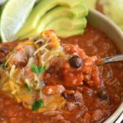 Vegetarian chili in a bowl with shredded cheese, lime, and avocado as garnish with spoon.
