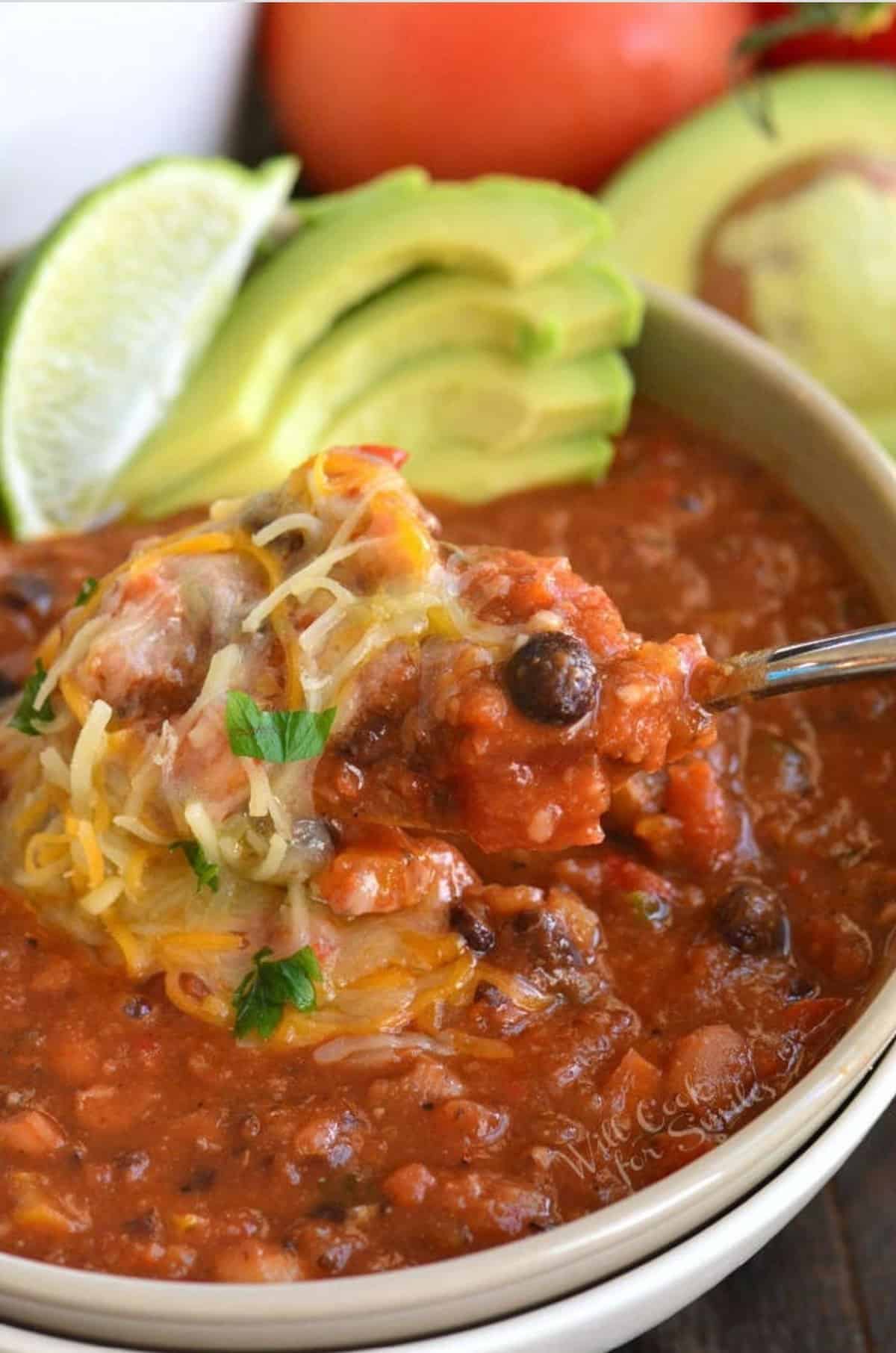 Vegetarian chili in a bowl with shredded cheese, lime, and avocado as garnish with spoon.