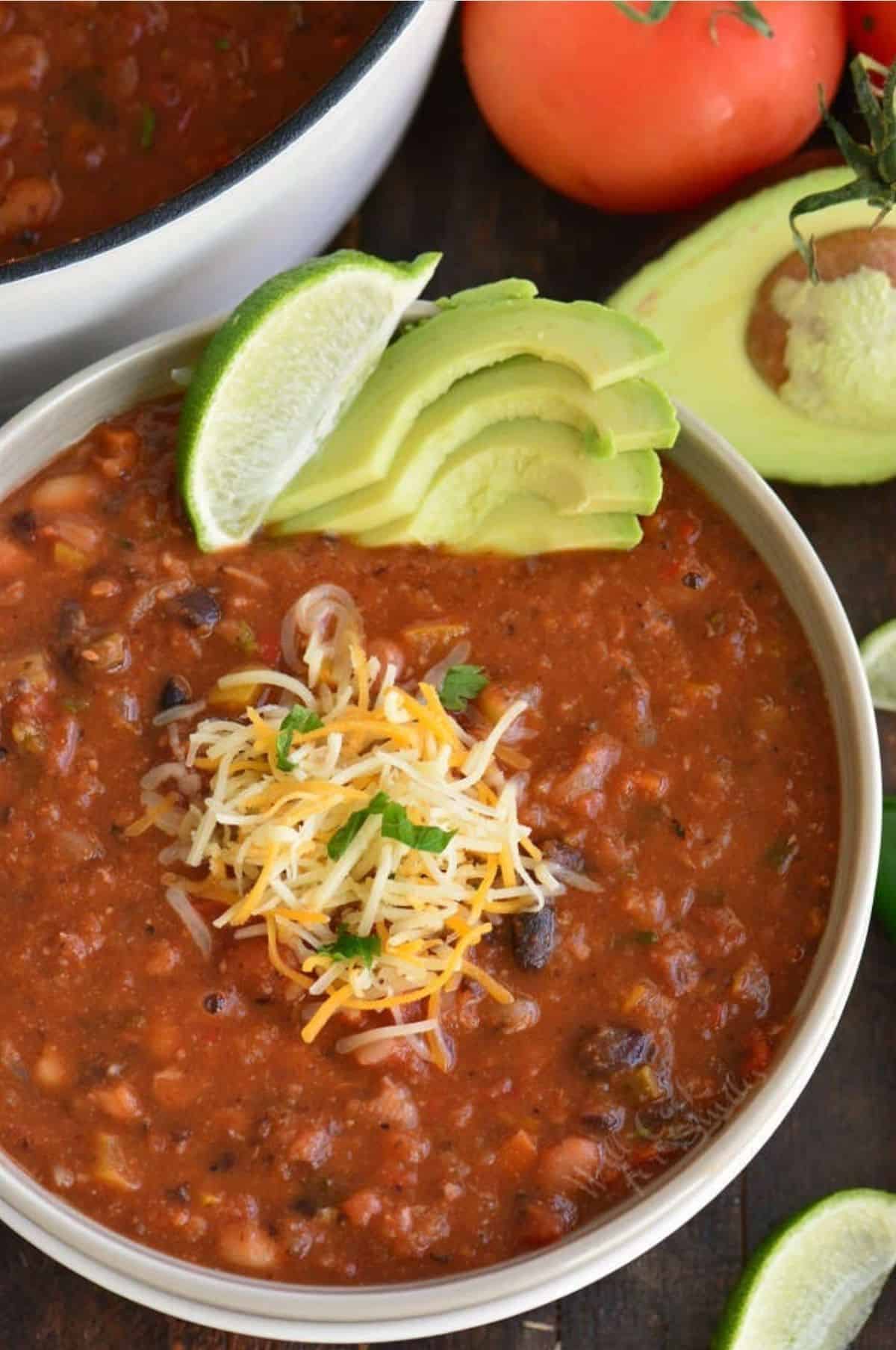 Vegetarian chili in a bowl with shredded cheese, lime, and avocado as garnish with tomato and avocado in the background.