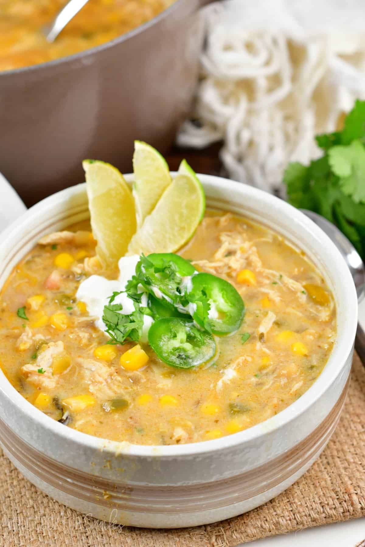 white chicken chili in a bowl with three sliced limes, jalapeno, and sour cream on top on a burlap placemat.