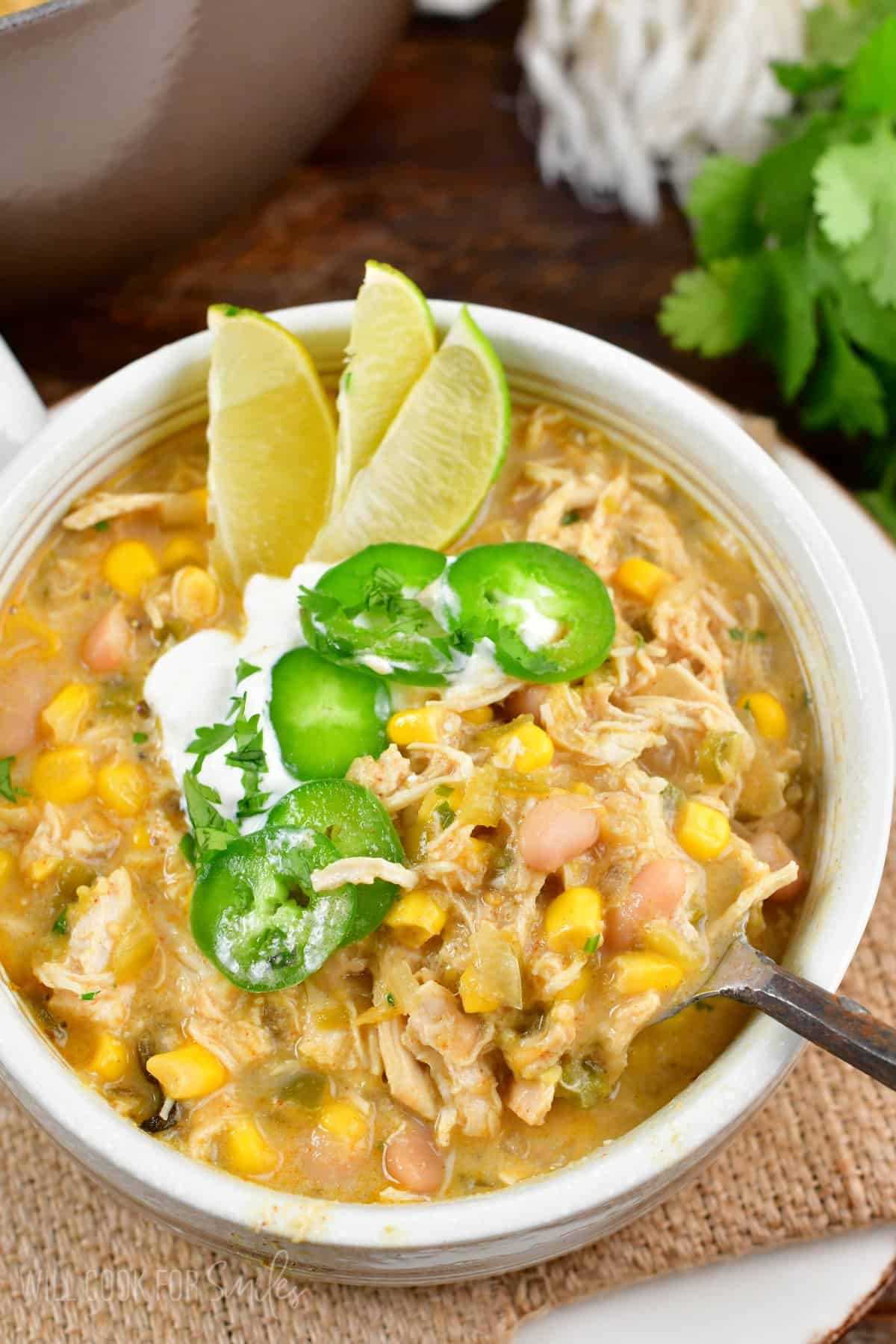 white chicken chili in a bowl with three sliced limes, jalapeno, and sour cream on top with a spoon scooping some out,