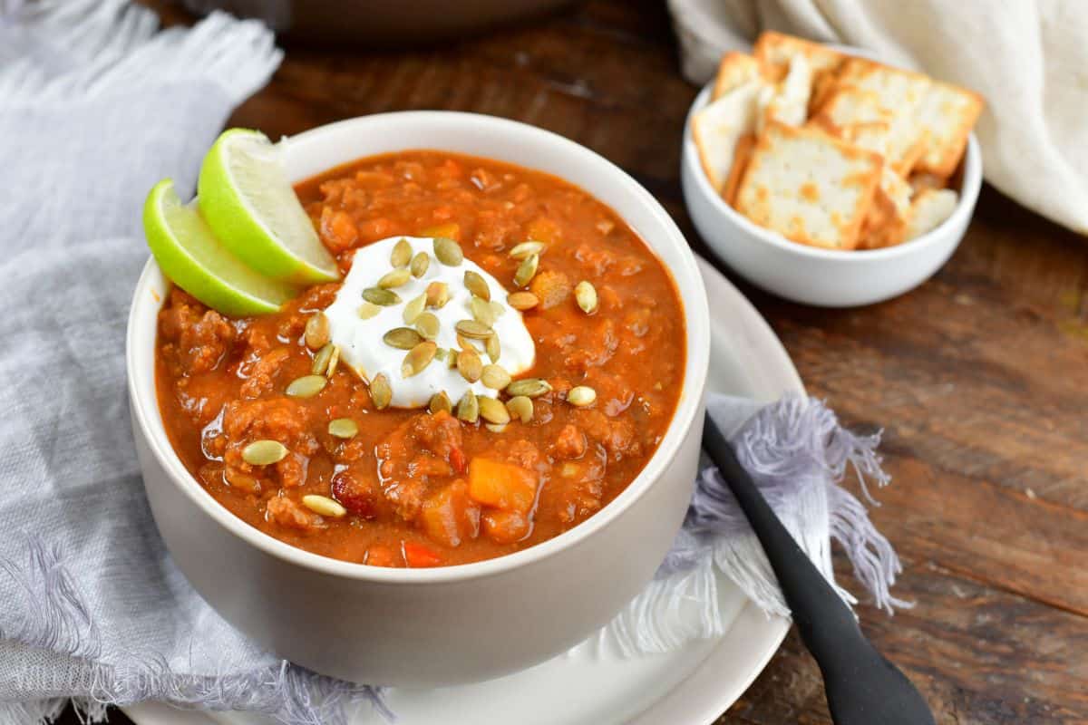 pumpkin chili in a bowl with a lime, sour cream, and pumpkin seeds as garnish on top on a plate with crackers in a bowl.