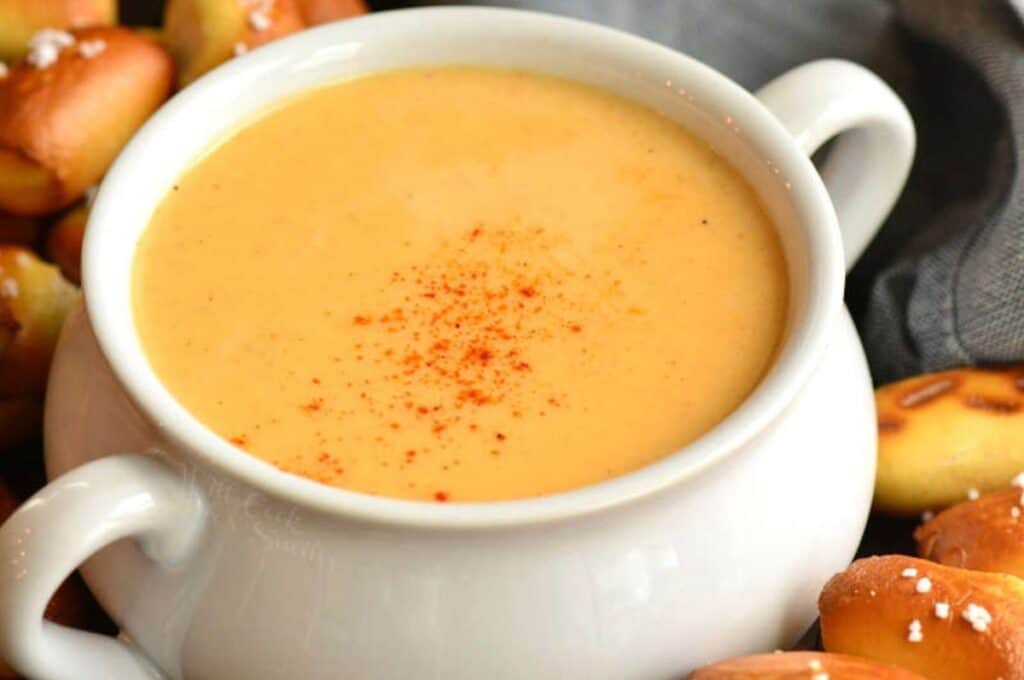 A white soup bowl filled with creamy cheese sauce, garnished with a sprinkle of paprika, is surrounded by pretzel bites. A gray cloth napkin is partially visible in the background.