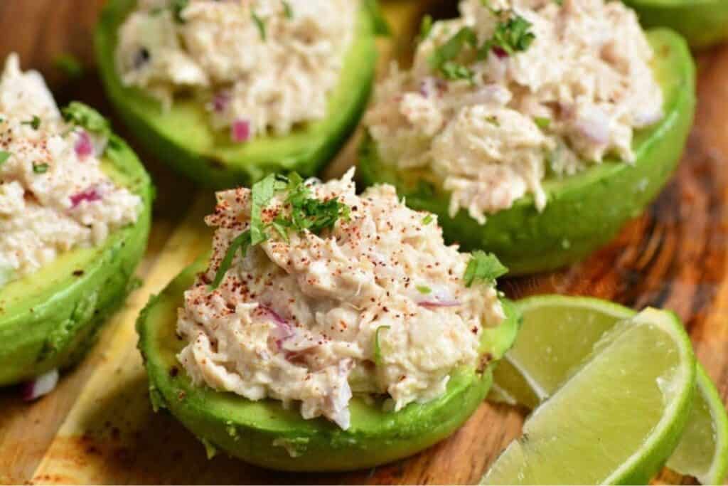 Half avocados filled with creamy chicken salad, garnished with chopped parsley and sprinkled with paprika, placed on a wooden board. Lime wedges are on the side.
