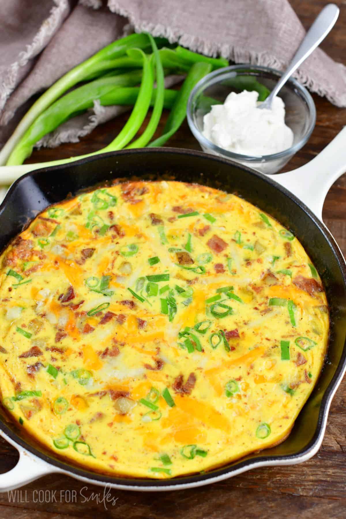 breakfast skillet topped with green onion and bacon next to sour cream and green onion.