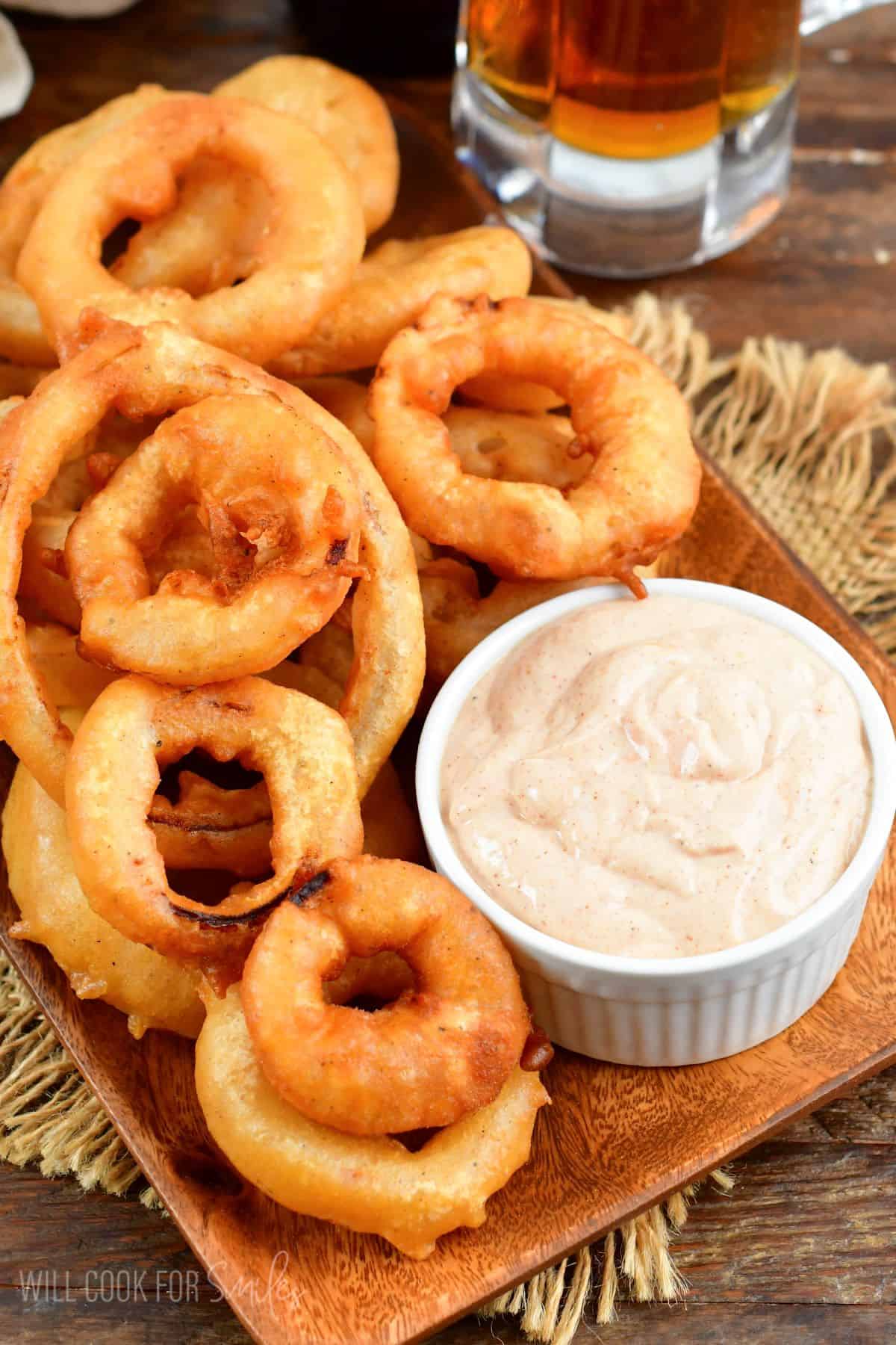 Beer battered onion rings on a wood plate with a dipping sauce.