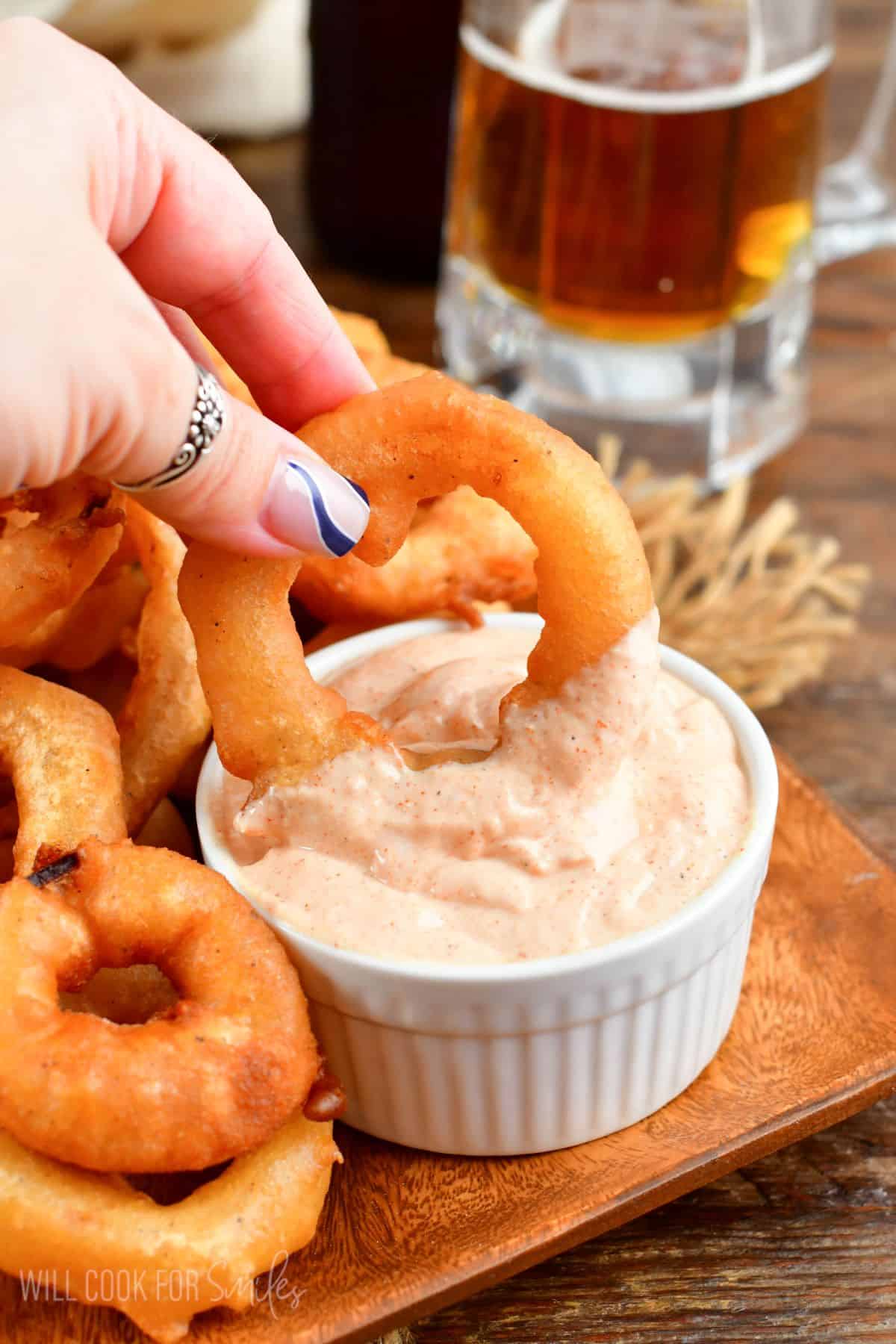 dipping a beer battered onion ring into sauce that is in a small bowl on a wood plate.