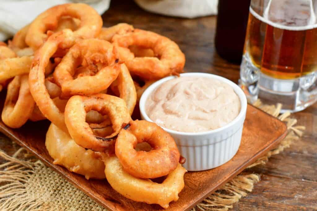A wooden tray filled with golden crispy onion rings sits next to a small white bowl of creamy dip. A glass of beer is visible in the background on a rustic wooden table.