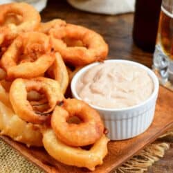 Beer Battered onion rings on a wood plate with a dipping sauce and a beer in the background.