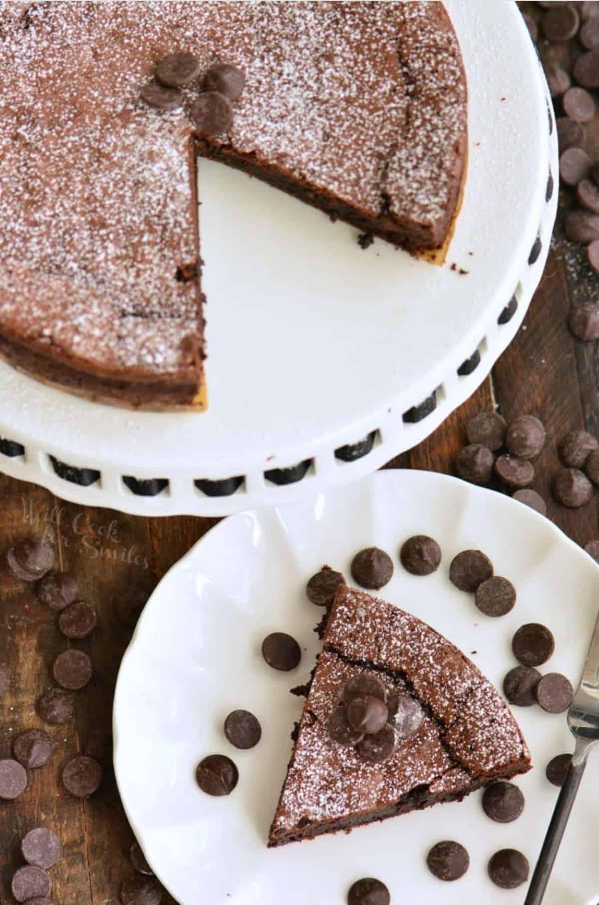 slice of chocolate cake on the plate next to the cake stand with the whole cake.