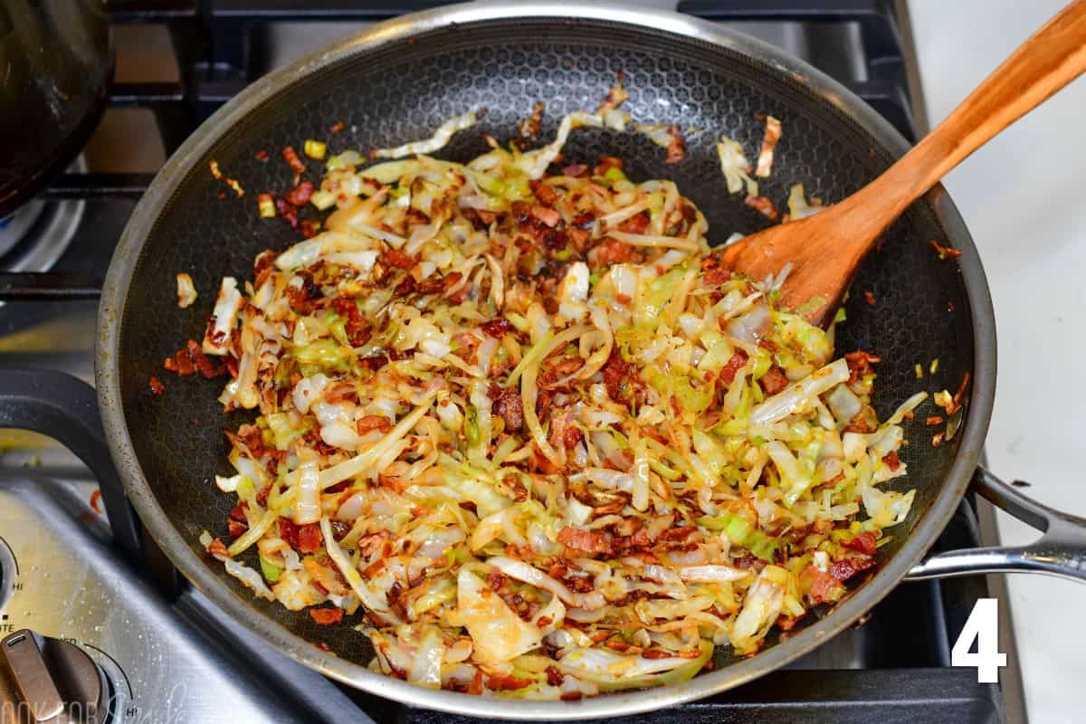 Cooking fried cabbage and bacon in a pan and stirring with a wooden spoon.
