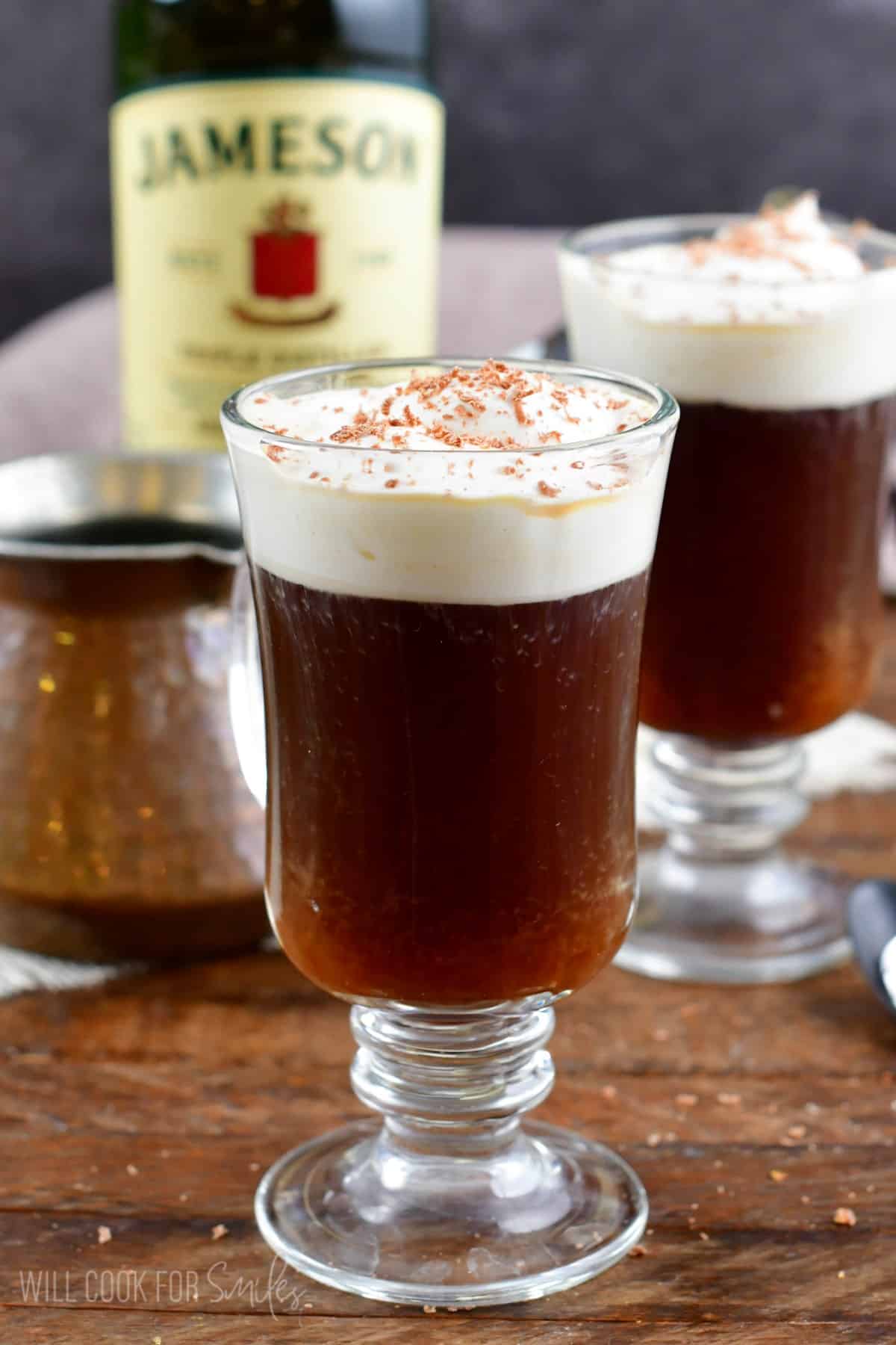 I glass filled with Irish coffee with another glass and a botle of Jameson in the background.