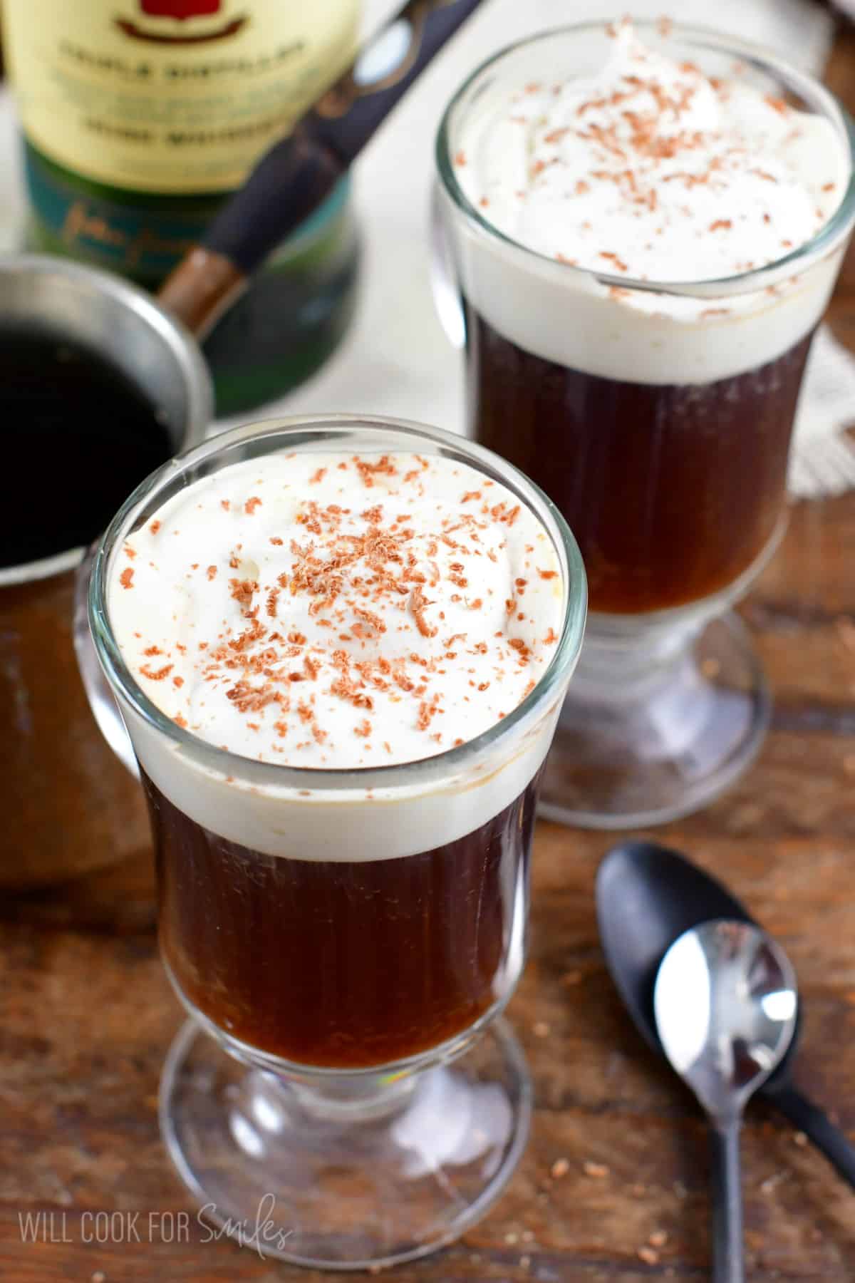 two cups of Irish coffee with cream on a wood surface with two spoons on the side.