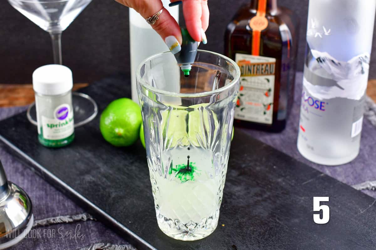 Adding green food coloring to a glass that is on a black wood surface with a bottle of vodka in tha background.