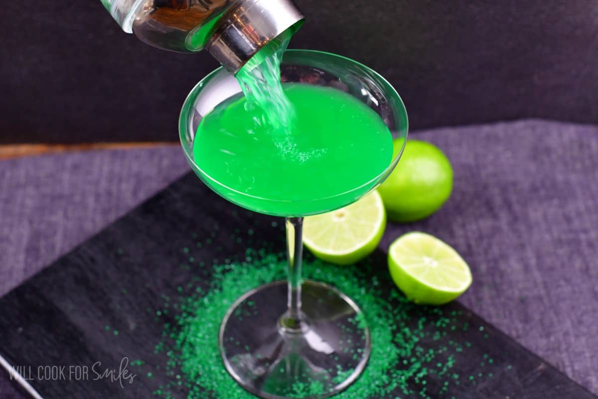 pouring the lime martini into a martini glass with green sprinkles around the stem of the glass and a lime sliced in half.