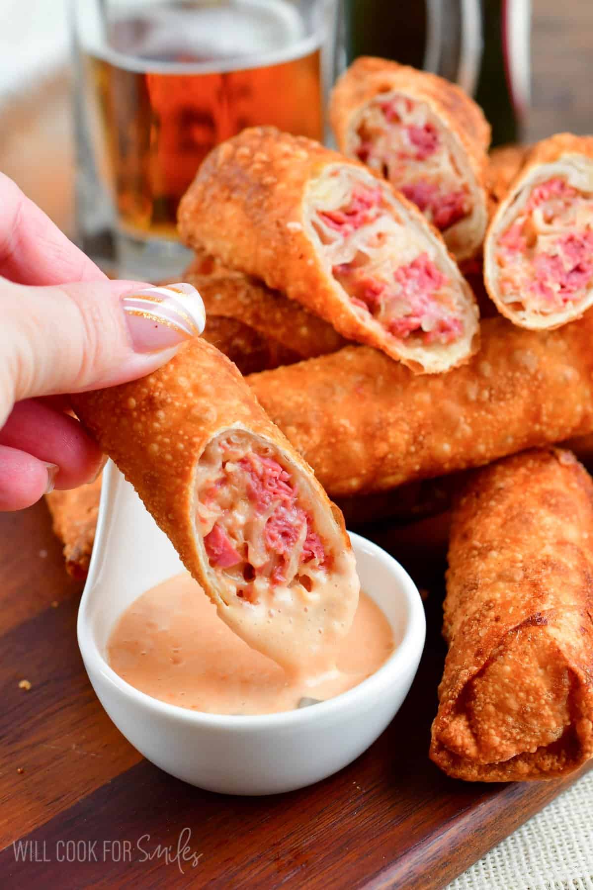 Dipping a reuben egg roll into dressing with the rest of the egg rolls on a wooden tray.