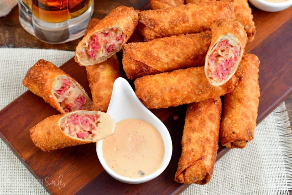 A wooden board with crispy, golden-brown egg rolls filled with a pinkish mixture, accompanied by a small white dipping sauce container. A glass of a dark beverage is partially visible in the background.