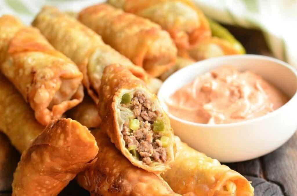 A close-up of several crispy egg rolls, one of which is cut in half to display a filling of ground meat and vegetables. A white bowl of creamy dipping sauce accompanies the egg rolls. The background is slightly blurred, accentuating the focus on the food.