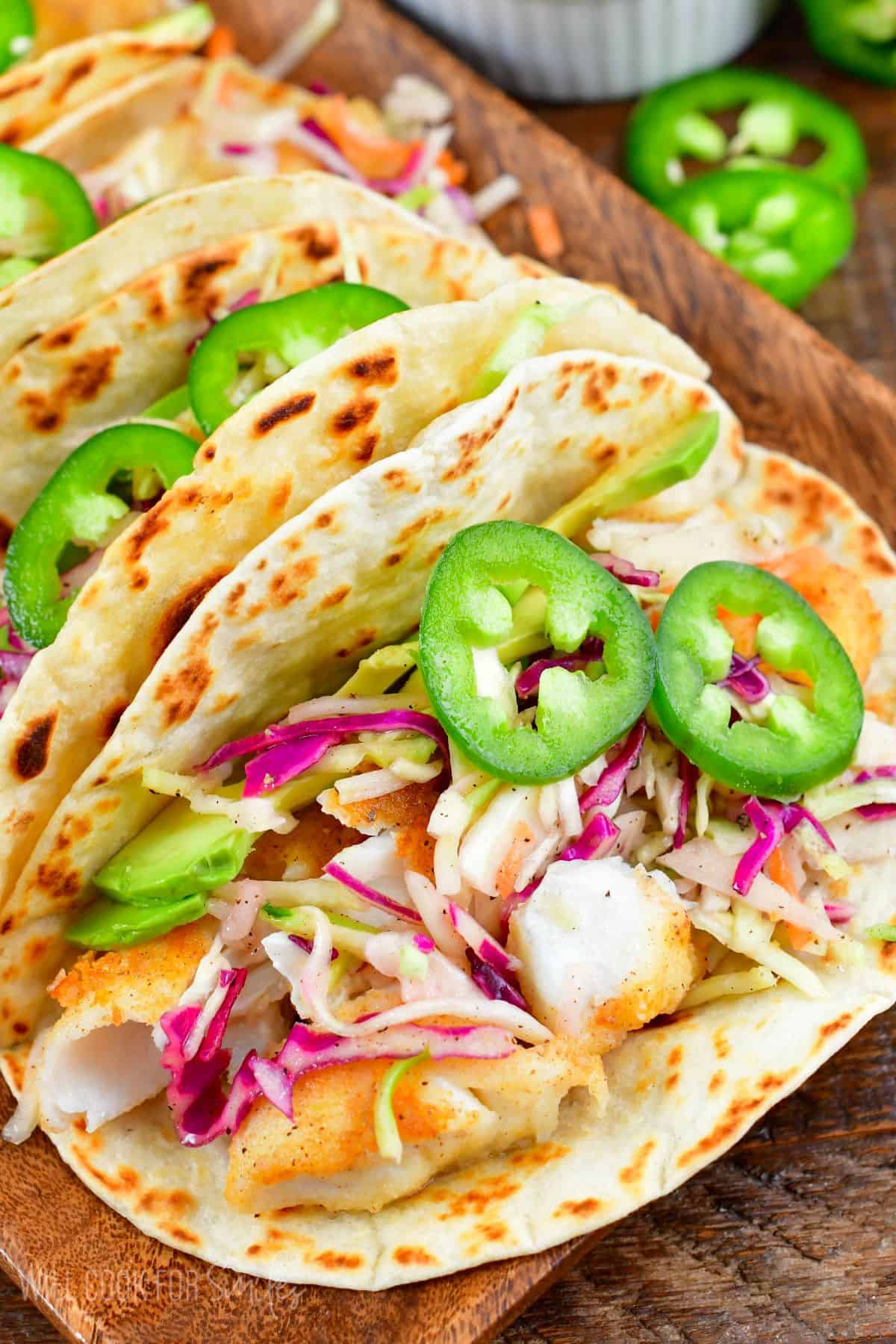 Fried fish tacos in flour tortilla with avocado, cabbage, jalapenos on top with a wood tray.