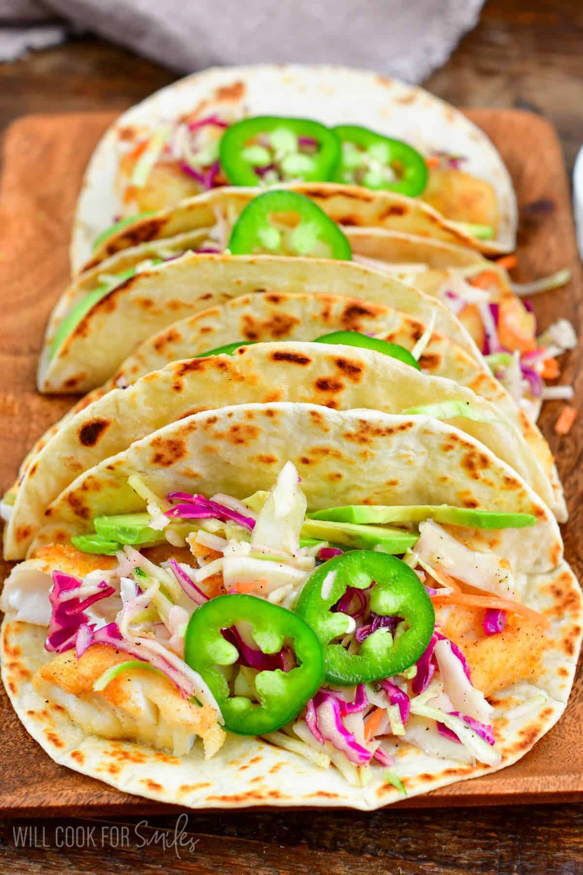 Four fish tacos lined up on a wood tray with cabbage, avocado, and jalapenos as topping.