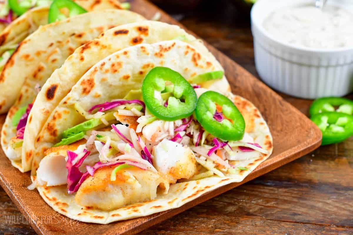 Fish tacos on a wood plate with cabbage, jalapeno, and avocado on a wood tray.