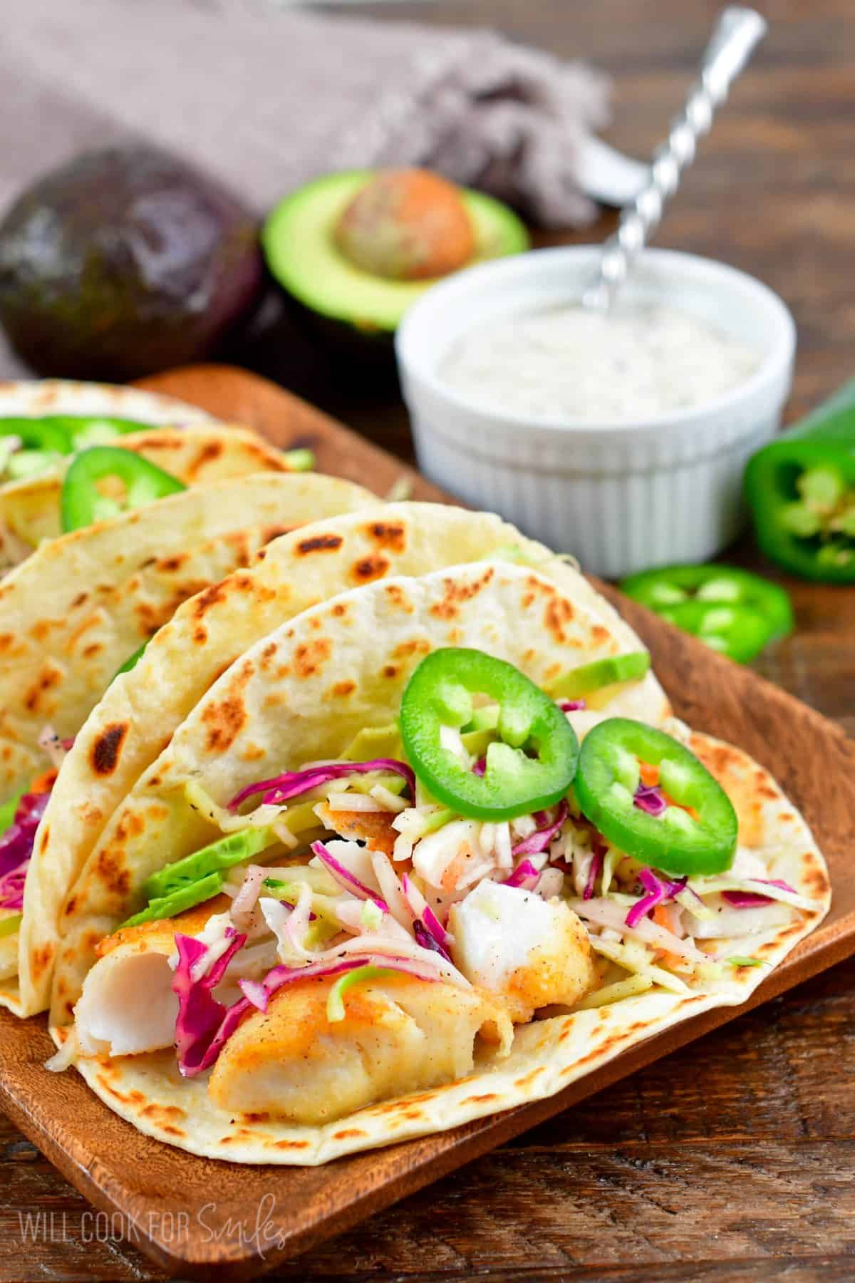 Fried fish tacos in a flour tortilla with jalapenos, cabbage, and avocado on a wood tray.