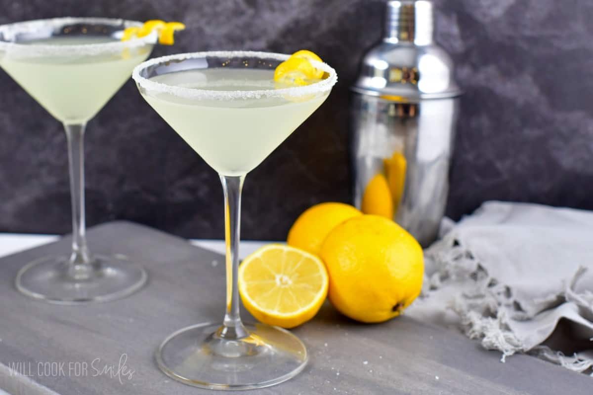 Lemon drop in a martini glass with a sugar rimmed glass and two whole lemons and one half of a lemon on a wood surface.
