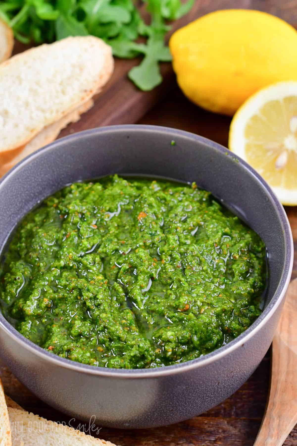 Lemon Pistachio Arugula Pesto in a bowl on a wood surface with a sliced lemon in the background.