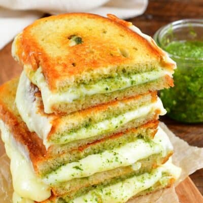 two pesto grilled cheese sandwiches cut in half and stacked up on a piece of parchment paper with a jar of pesto to the right.