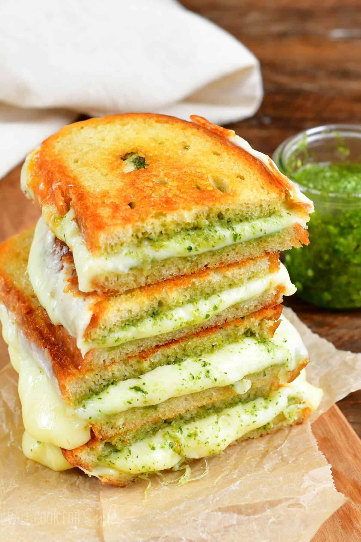 two pesto grilled cheese sandwiches cut in half and stacked up on a piece of parchment paper with a jar of pesto to the right.