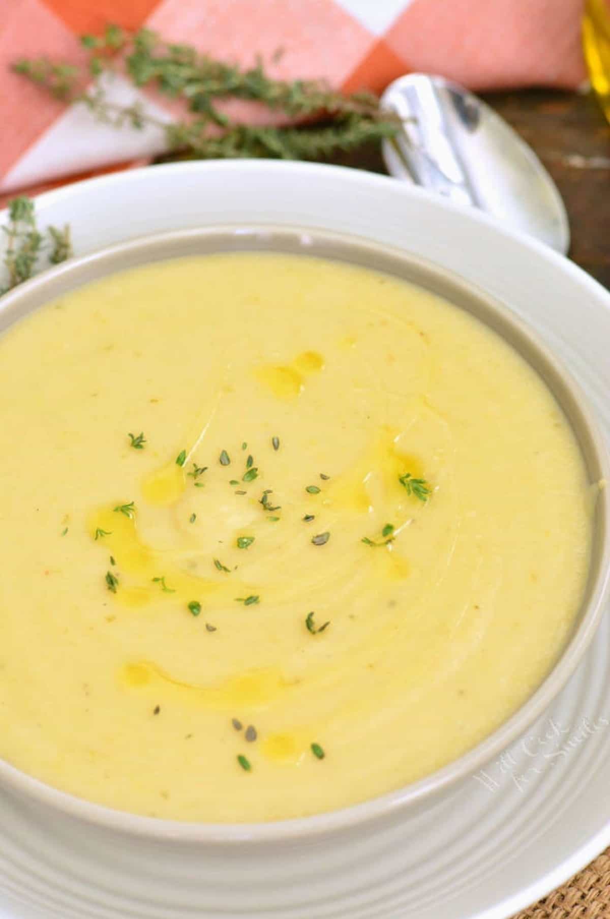 Potato leek soup in a bowl on top of a plate with rosemary sprinkled on top as garnish.