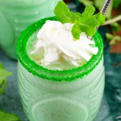 Shamrock shake in a glass with sprinkles on the rim of the glass and whipped cream and mint as garnish.