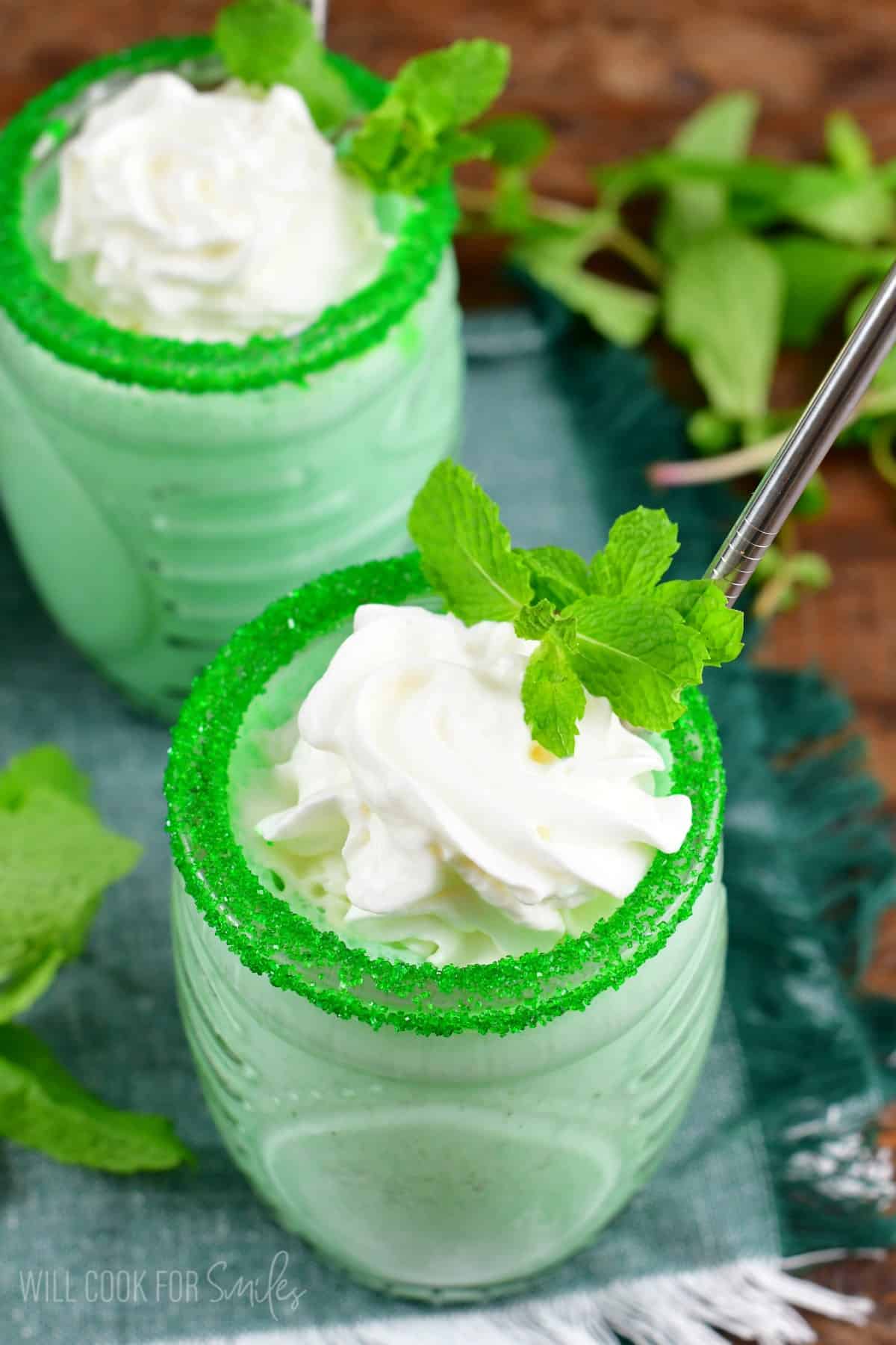 two shamrock shakes in a glass with green sprinkles on the rim, whipped cream and a piece of mint as garnish.
