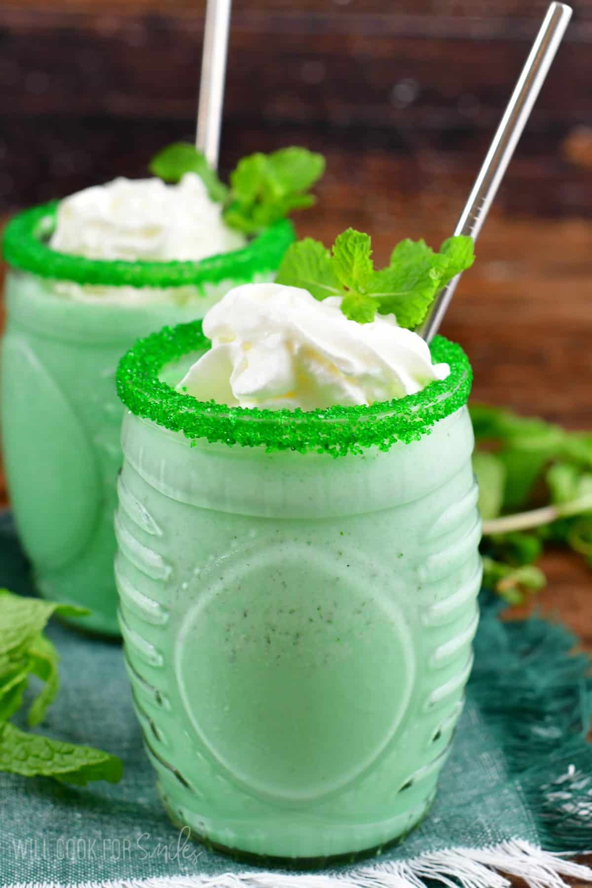 Green shamrock milkshake in a glass with whipped cream. sprinkles on rim of glass, and a metal straw.