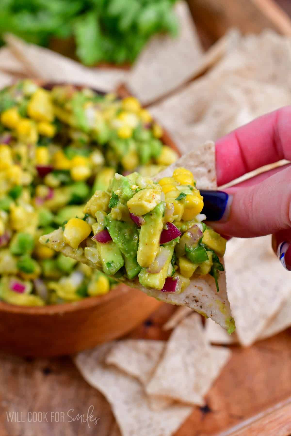 Holding a chip with avocado salsa on top and the bowl of avocado salsa in the backgound.