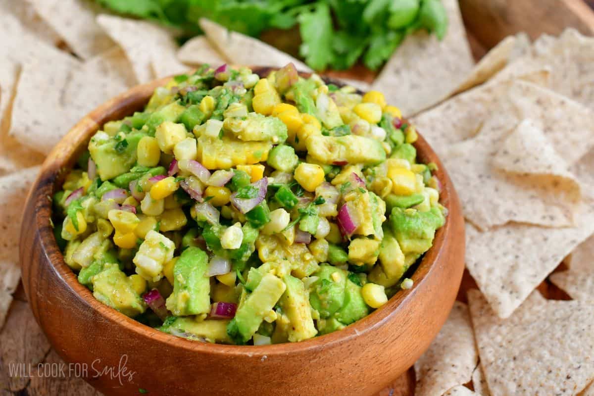 Avocado Salsa in a wood bowl on top of a wood tray and tortilla chips around the tray.