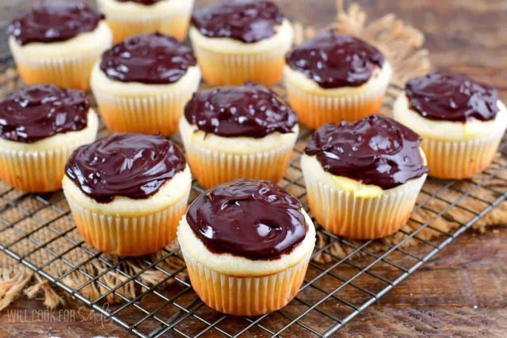 A dozen vanilla cupcakes topped with glossy chocolate frosting are arranged on a metal cooling rack. The cupcakes are placed on a wooden surface with a rustic fabric accent underneath.