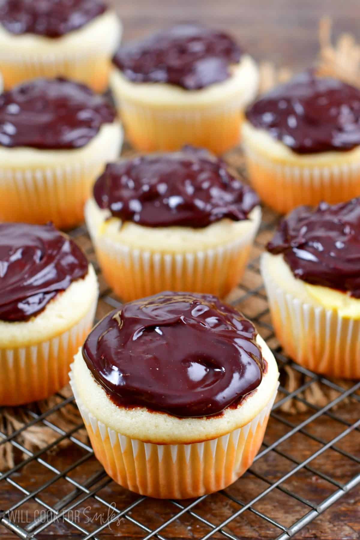 Boston Cream cupcakes on a wire rack with chocolate frosting on top on a wood surface.