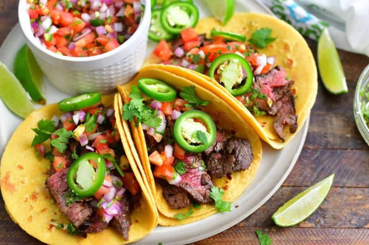 three carne asada tacos on a plate with toppings, limes, and pico de gallos.