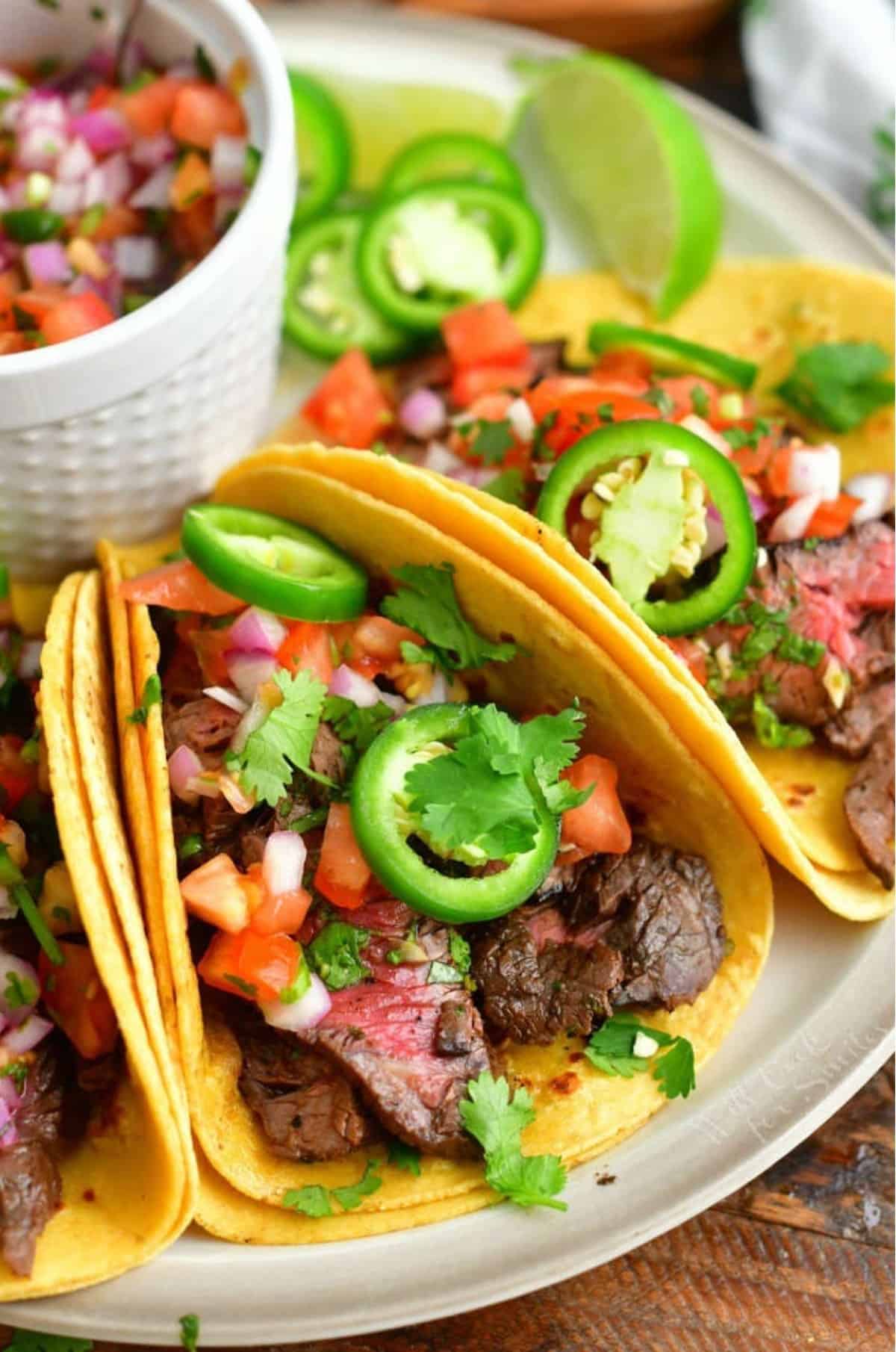 carne asada tacos topped with pico de gallo and jalapenos on a plate.