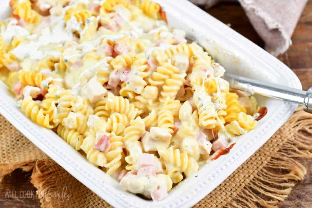 A white rectangular dish filled with creamy chicken and bacon ranch pasta featuring spiral noodles, chunks of chicken, and pieces of bacon, topped with a creamy white sauce. A serving spoon rests in the dish. The dish is set on a beige cloth.