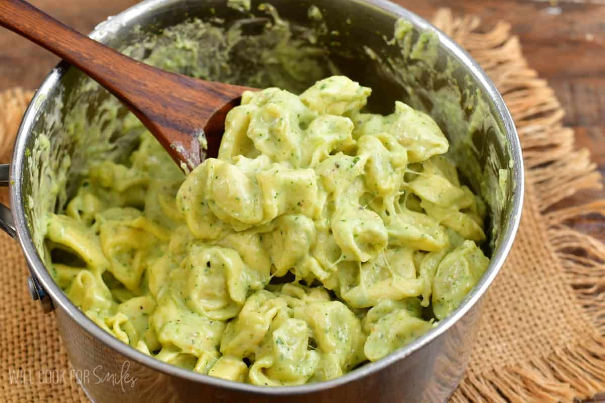 scooping pesto alfredo tortellini out of the pan with a wooden spoon.