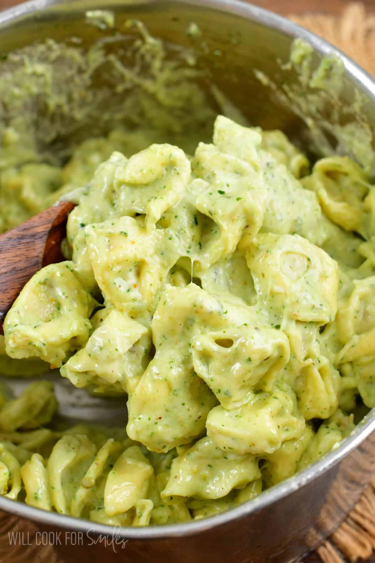 Using a wooden spoon to scoop up some of the pesto alfredo tortellini in a pot.