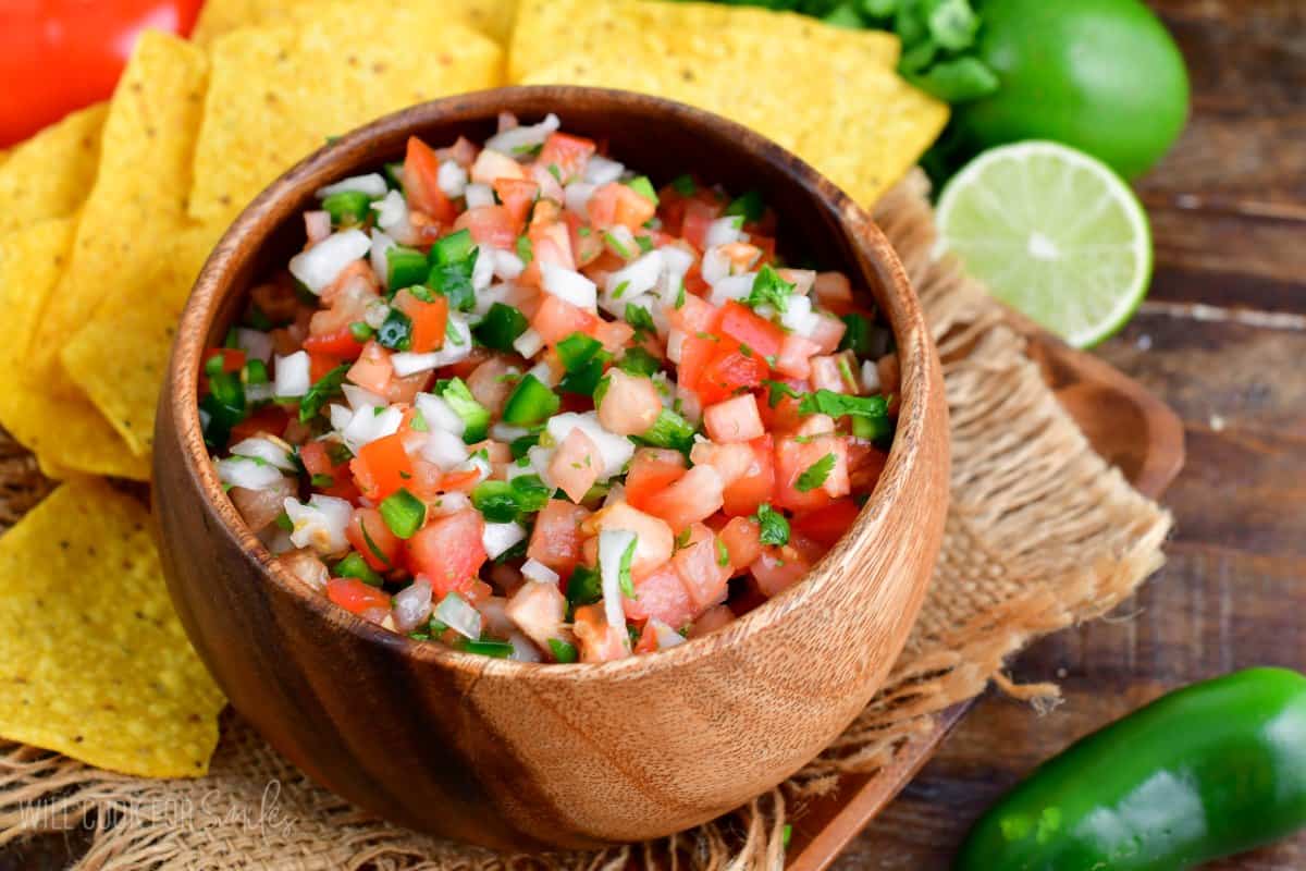 Pico de Gallo in a bowl on a wood plate with tortilla chips and a lime and jalapeno to the side.